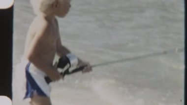 Bir çocuğun elindeki oltayla yavaşça sallanırken, okyanus dalgaları dalgaları dalgalanırken kıyıda durduğu klasik bir fotoğraf.