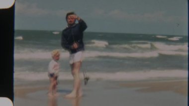 Genç oğlunun oltasını ayarlayan bir babanın klasik çekimi, arka planda okyanus dalgaları çarpıyor, kıyıda samimi bir yakınlaşma anı yakalıyor.