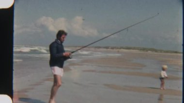 Bir baba ve oğlunun, sakin bir sahilde birlikte olta atarken, okyanus kıyısında huzurlu ve neşeli bir bağ anının tadını çıkarırken çekilmiş klasik bir fotoğrafı.