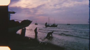 İki adam arka planda yelkenli teknelerle deniz kıyısında su topluyor, sakin bir kıyı manzarasına karşı hazırlanıyorlar.