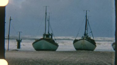 Sahile demir atmış yelkenli tekneler, arka planda dramatik bulutlu bir ufuk ve dalgalarla yavaşça sallanıyordu.