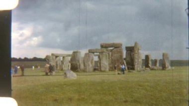 Turistlerin yemyeşil bir alanda arka planda yükselen görkemli Stonehenge sütunlarıyla dolaşarak tarih ve merak hissini yakaladıkları klasik bir fotoğraf.