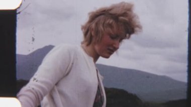 8mm 'lik bir görüntü, arka planda görkemli dağlar tarafından çerçevelenmiş sakin bir su kütlesiyle çölde gezinen bir bayanın görüntüsü.