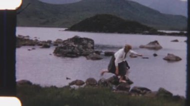 Kardeşi yakındaki bir kayadan yakından izlerken bir bayanın sahildeki kayaları dikkatle gezdiği 8 mm 'lik bir görüntü.