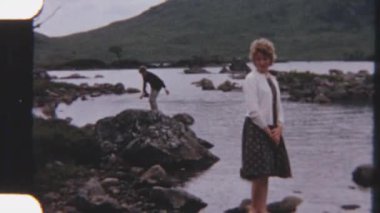 8 mm 'lik bir görüntü, sakin suların üzerindeki bir kayanın üzerinde duran bir bayanın görüntüsü, bereketli doğa ve dağlarla çevrili.