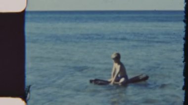 Kaygısız bir çocuk, sakin okyanus sularında bir şamandıraya biniyor, 8mm 'lik klasik bir filme çekiliyor, dingin dalgalar etrafında yavaşça dalgalanırken.