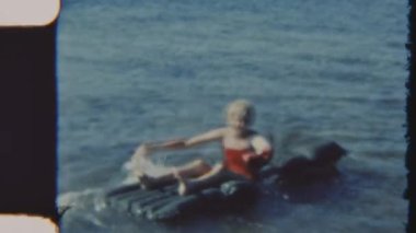 Kırmızı mayo giymiş bir kız güneşli bir günde sakin okyanus sularında yüzerken eliyle kürek çekiyor ve klasik bir 8 mm 'lik filmle çekiliyor.