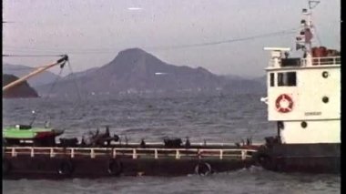 Kasvetli, eski bir VHS videosu, güvertesi çeşitli yapı ve ekipmanlarla dolu bulutlu gökyüzü altında uzak dağları olan bulanık bir sahil şeridine kurulmuş bir gemiyi gösteriyor.
