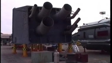 Bir kamera, sol tarafta büyük metal bir yapı, muhtemelen bir geminin egzoz veya havalandırma sisteminin parçası olan silindirik metal boruları olan bir tersanenin çalışma alanını yakalar.