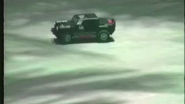 Düşük çözünürlüklü, analog bir video, kırmızı aksanlı siyah uzaktan kumandalı bir oyuncak arabayı gösteriyor. Sert bir off-road tarzında dizayn edilmiş, kumlu ve desenli zeminde sürüyor.
