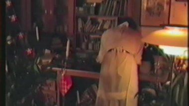 Eski bir ev videosundan bir sahne Noel arifesinde bir çocuk ve küçük bir çocuk Noel ağacının etrafında yürürken yetişkin bir kadının rahat, şenlikli bir oturma odasında durduğunu gösteriyor.