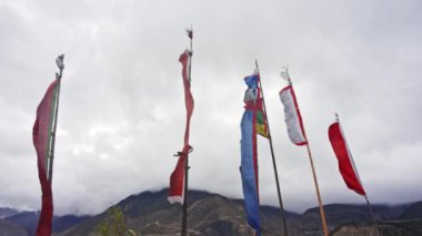 Rüzgarda dalgalanan renkli dua bayrakları dramatik bulutlu gökyüzüne karşı, arka planda yükselen görkemli Himalaya dağ sıraları, dingin ve ruhani bir atmosfer yaratıyor.