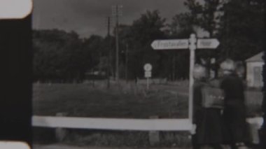 8mm 'lik bir filmde çekilen nostaljik siyah-beyaz sahne, kırsal yoldaki bir levhanın yanından geçen klasik giysili bir aileyi gösteriyor, geçmişten gelen zamansız ve huzurlu bir anı anımsatıyor.