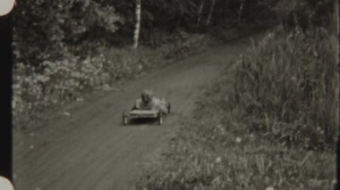 8 mm 'lik filmle çekilen, ağaçlarla çevrili, bir sabun kutusunun içinde, orman yolunda yuvarlanan genç bir çocuk.