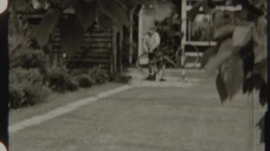 8mm 'lik siyah beyaz bir fotoğraf, evinin yanında bisikletinin lastiğiyle ilgilenen genç bir çocuk.