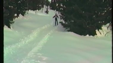 Genç bir kayakçı, arkasında küçük yeşil bir kızakla karla kaplı bir yamaçtan aşağı süzülür ve onu daha genç bir kayakçı takip eder.