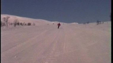 Analog bir video, kar yamacından kameraya doğru süzülen kırmızı ceketli bir kayakçıyı, ağaçlardan oluşan bir kış manzarasına ve hafif kumlu bir kalite ile bulutlu bir gökyüzüne karşı çeker.