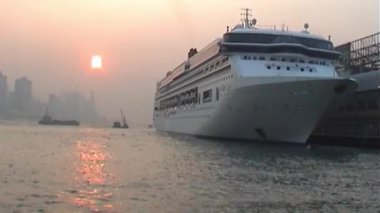 Büyük bir yolcu gemisi Hong Kong 'da bir limana yanaştı. Güneş, sisli ufuk çizgisi, yüksek binalar ve arka planda hareketli liman aktiviteleriyle, yumuşak turuncu bir parıltı salıyor.