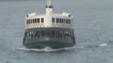 Yakın plan çekimde sakin ve berrak sulardan geçen bir feribot görülüyor. Hareket ederken küçük dalgalar yaratıyor. Arka planda binalar ve diğer yapılar görünüyor.