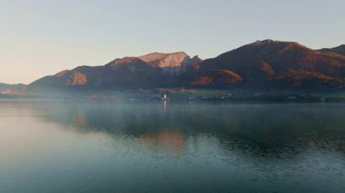 Bir dron, heybetli dağlar ve yansıtan sularla çevrili sakin bir göl kenarı köyüne sorunsuzca yaklaşıyor.