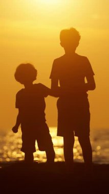 Ilık altın ışık suyu yansıtırken, iki çocuk canlı bir günbatımına karşı gölgelenmiş kayaları denize atıyorlardı.