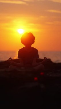 Güneş batarken kayalık bir kıyıda Sukhasana pozu veren sakin bir çocuğun silueti huzurlu ve huzurlu bir atmosfer yaratıyor.