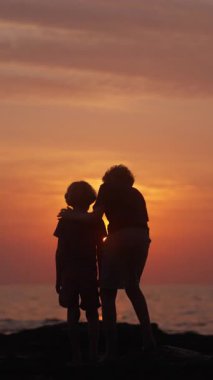 Siluet içindeki iki kardeş, sahilde huzurlu bir anı paylaşıyor, günbatımını sessizce seyrediyorlar.