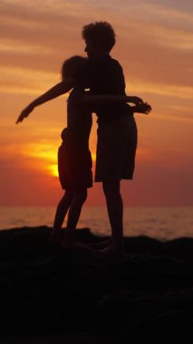Silüet içindeki iki kardeş, çarpıcı bir okyanus günbatımına karşı yürekten bir kucaklaşmayı paylaşıyor. Derin bir bağ ve duygu anı yakalıyorlar.