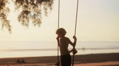 Bir çocuk salıncakta duruyor, güneş suyun üzerinde yavaşça süzülüyor, sakin sahnenin üzerine altın bir parıltı saçıyor