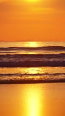 Gün batımının ılık parıltısının altında süzülen yumuşak dalgalarla sakin bir sahil sahnesi.