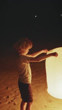 Bir çocuk gece plajda durur, sıcak ışığı karanlığı aydınlatırken parlayan bir fener tutar.