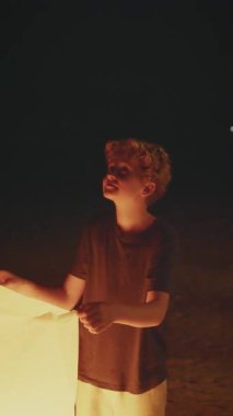 Genç bir çocuğun yüzü, gece sahilde durduğu sırada bir fenerin sıcak ışığında usulca parlıyor. Huzurlu ve büyüleyici bir sahne yaratıyor.