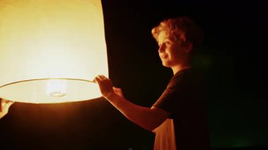 Genç bir çocuk gece gökyüzünün altında duruyor. Elinde parlayan bir fener var ve onu karanlığa salmaya hazırlanıyor.