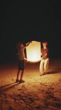 Çocuklar geceleri plajda durup, sıcak ışığı karanlığa doğru sürüklenirken gökyüzüne parlayan bir fener salıyor.