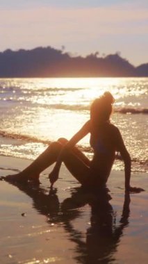 Siluetli bir kız kumsalda kumlara resim çiziyor, güneşli bir okyanus arkaplanı ile sıcak ve huzurlu bir manzara yaratıyor..