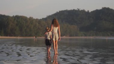 Çocuklar kıyı boyunca yalınayak yürürler, arkadan bakarlar, yemyeşil tepeler ve arka planda sakin doğal bir manzara