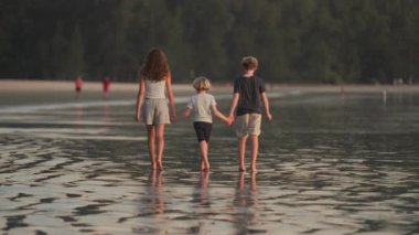 Arka planda, arka planda tropik ağaçlarla ıslak sahil kumu üzerinde çıplak ayakla yürüyen çocuklar var.