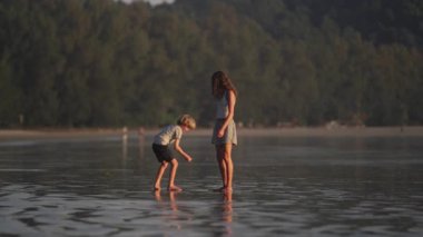 Çocuklar ıslak kumsalı eğlenceli bir şekilde keşfediyor, yoğun yeşillik arkalarında yemyeşil bir zemin oluşturuyor.