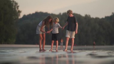 Islak kumda çıplak ayakla yürüyen çocuklar, yemyeşil ağaçlar tarafından çerçevelenmiş, kıyı şeridinin huzurlu güzelliğinin tadını çıkarıyorlar.