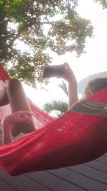 Bir kız, huzurlu ve huzurlu bir manzaranın ortasında, bir hamakta, akıllı telefonuyla doğanın güzelliğini yakalıyor.