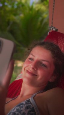 Genç bir kız kırmızı bir hamakta uzanır, akıllı telefonuyla kayar ve doğada huzurlu bir anın tadını çıkarır.