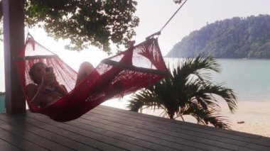 Genç bir delikanlı kırmızı bir hamakta rahatlar, akıllı telefonunda yavaşça sallanır.