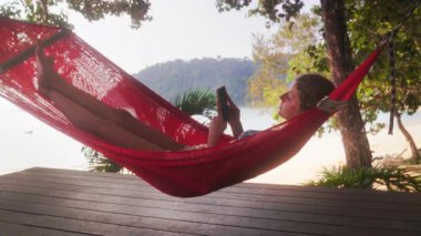 Genç bir kız açık havada bir hamakta uzanıyor, akıllı telefonunu kullanarak huzurlu bir ortamın tadını çıkarıyor.