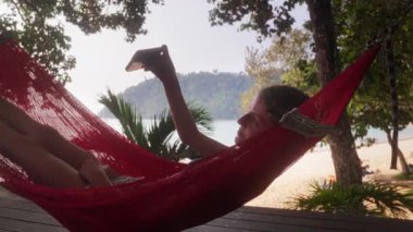 Neşeli bir genç, güneşli bir günde kırmızı bir hamakta uzanırken gülümser ve selfie çeker.