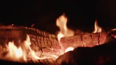 Kamp ateşinde yanan odunların yakın plan çekimi. Parlayan alevler karanlıkta yanıp sönüyor ve çatırdıyor. Sıcak ve davetkar bir atmosfer yaratıyor.