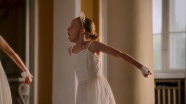 Ağır çekimde, parlak detaylar ve taclarla süslenmiş akıcı beyaz elbiseler içinde kollarını zarif bir şekilde uzatan genç dansçılar görülüyor.
