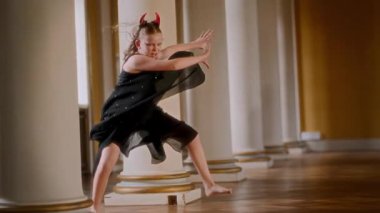 Genç kız, gösterişli salonda, uzun, zarif sütunlarıyla, dalgalı siyah elbiseler ve kırmızı şeytan boynuzlarıyla, etkileyici el hareketleriyle dramatik bir poz verirken, çağdaş dans gösterisi sergiliyor.