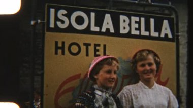 Isola Bella Otel tabelasının önünde gülümseyen ve poz veren iki genç kızın İtalyan maceralarının neşeli bir anını yakaladıkları 8 mm 'lik bir görüntü.