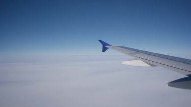 Uçağın içinden hava manzarası, kabarık bulutların üzerinde süzülüyor, arka planda canlı mavi bir gökyüzü, uçak kanadı tarafından çerçevelenmiş.
