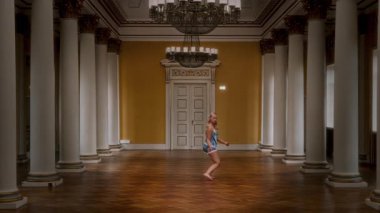 Gecelik giyen çıplak ayaklı genç bir kızı gösteren statik bir çekim. Büyük bir salonda akıcı bir şekilde hareket ediyor. Yüksek sütunlarla çevrili.